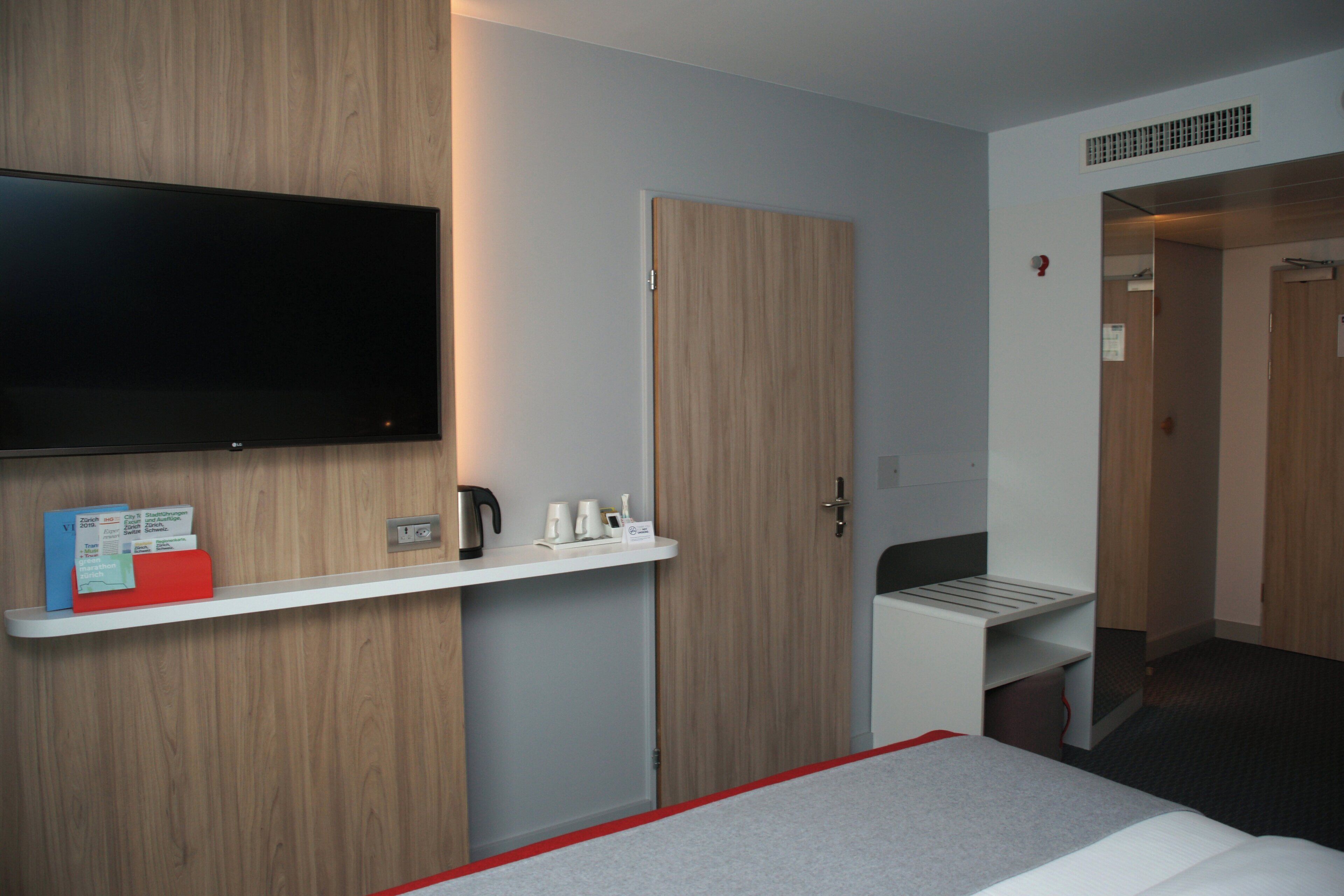 Habitación de hotel con televisor de pantalla plana montado en la pared, una mesita con hervidor y tazas, una carpeta roja y una puerta que da a otra habitación.