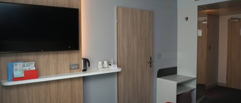 Habitación de hotel con televisor de pantalla plana montado en la pared, una mesita con hervidor y tazas, una carpeta roja y una puerta que da a otra habitación.