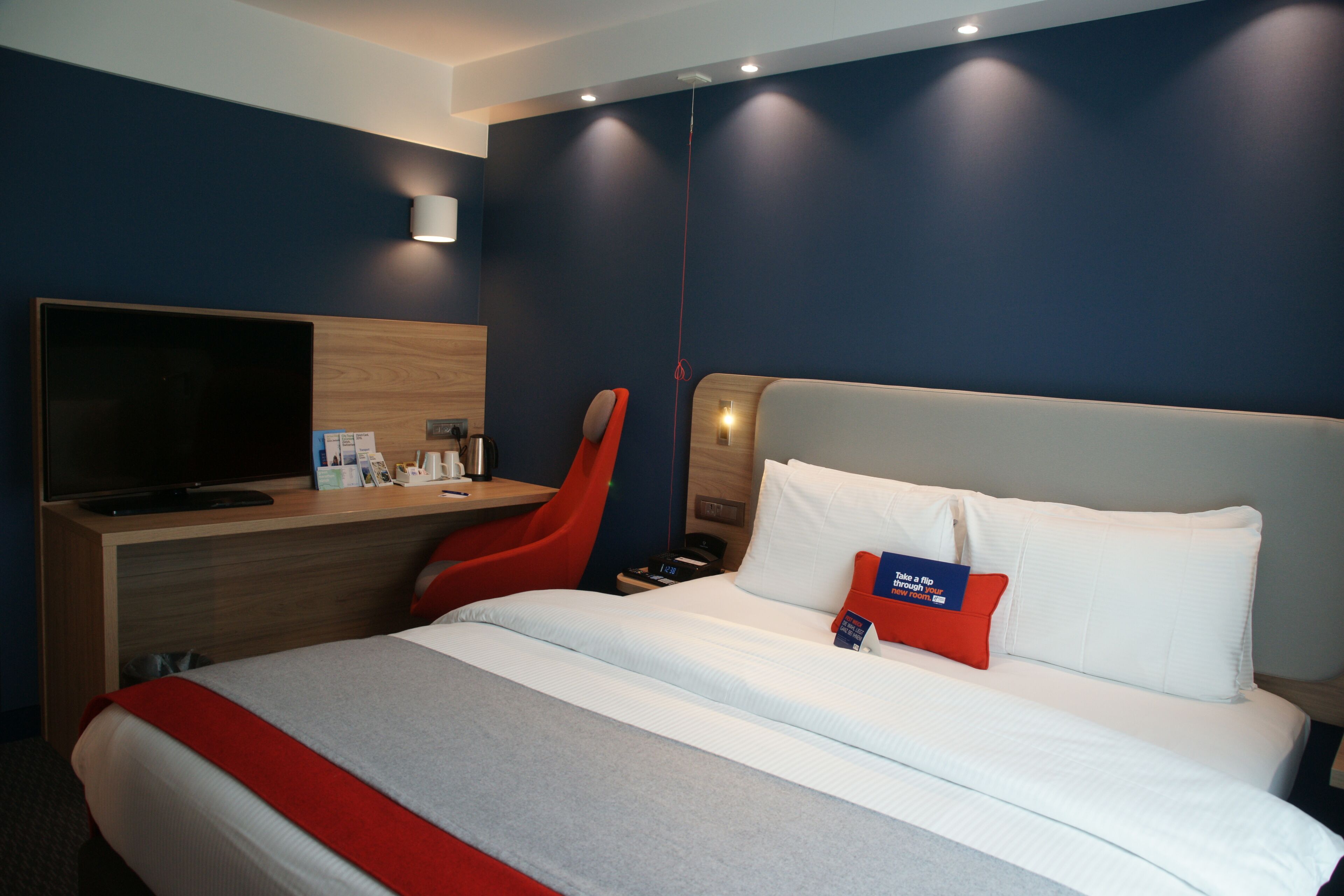 A modern hotel room with a large bed, a desk with a flat-screen TV, a red chair, and a nightstand.