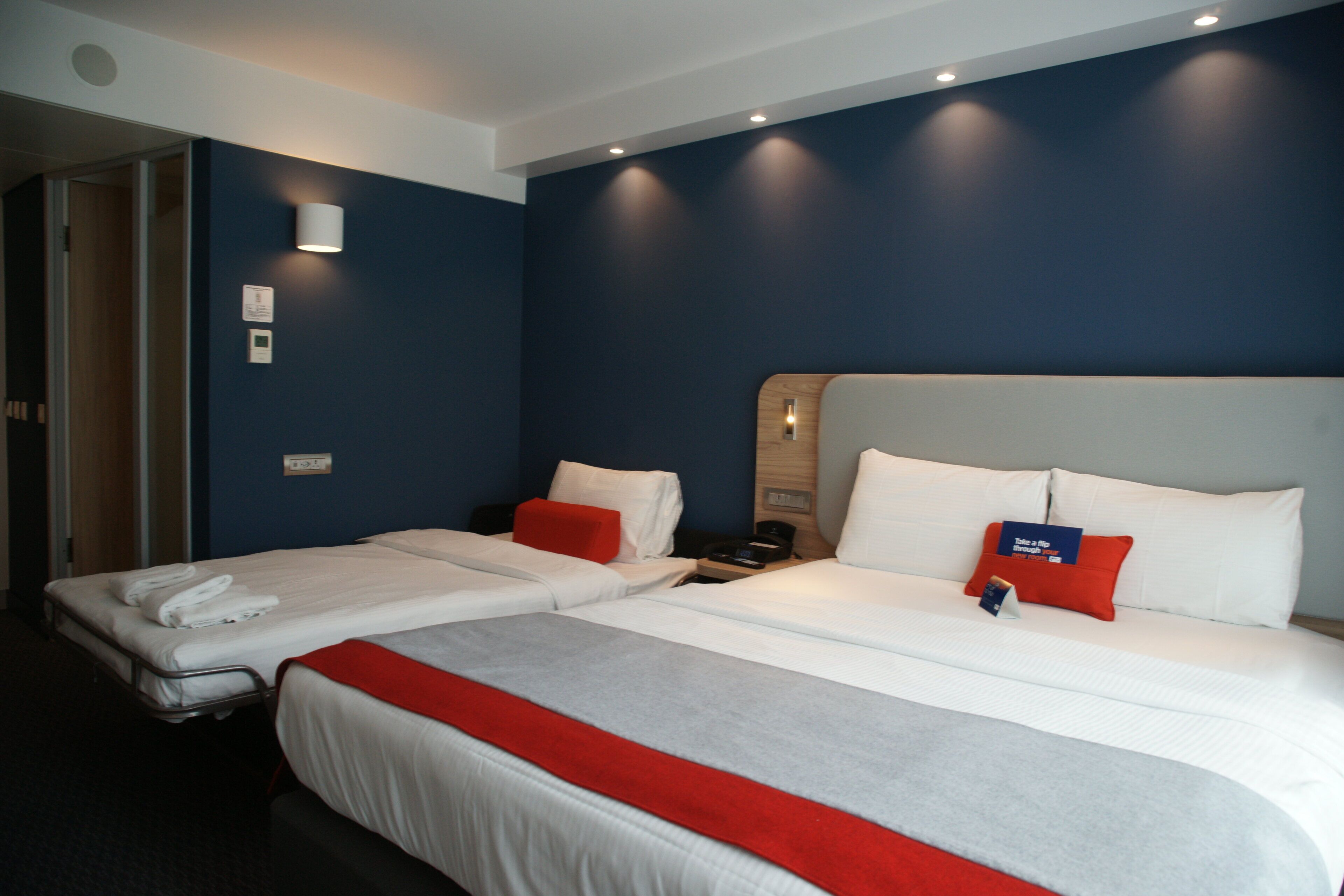 Habitación de hotel con dos camas, cada una con ropa blanca y detalles rojos, una mesita de noche con lámpara y un termostato en la pared.