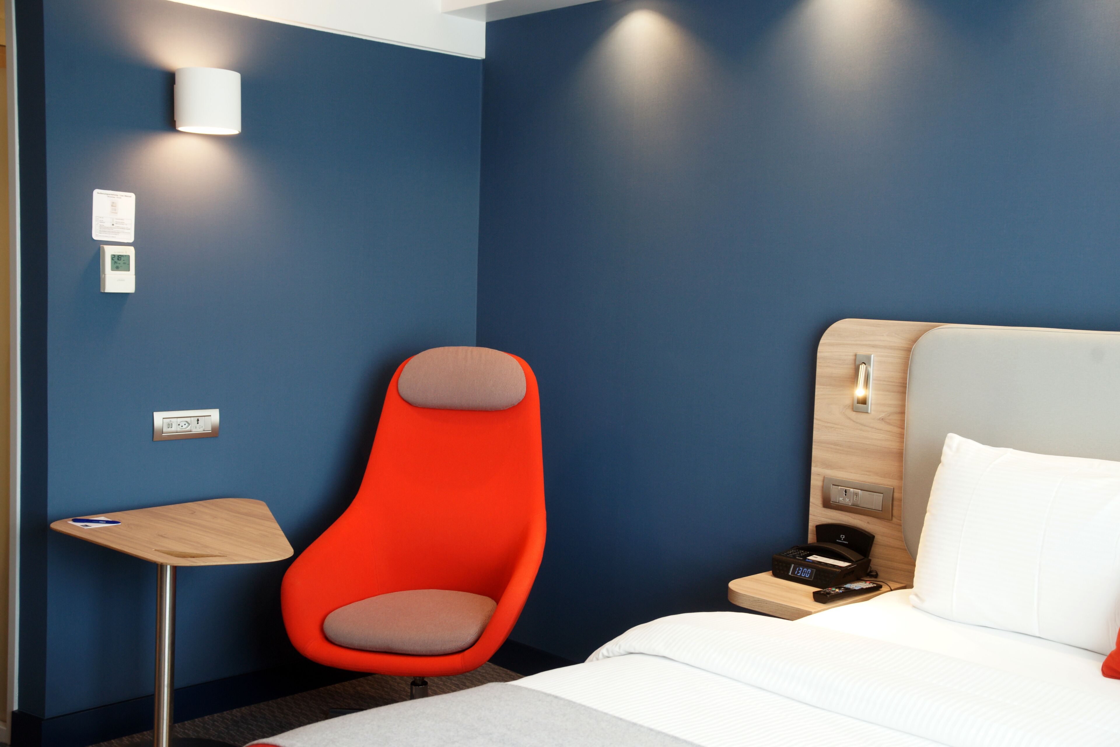 A hotel room with a bed, a wooden nightstand, a red chair, a small table, and a wall-mounted light fixture.