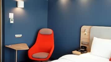 A hotel room with a bed, a wooden nightstand, a red chair, a small table, and a wall-mounted light fixture.