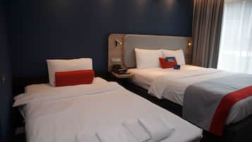Habitación de hotel con dos camas, cada una con ropa blanca y detalles rojos, una mesita de noche con teléfono y una ventana con cortinas.