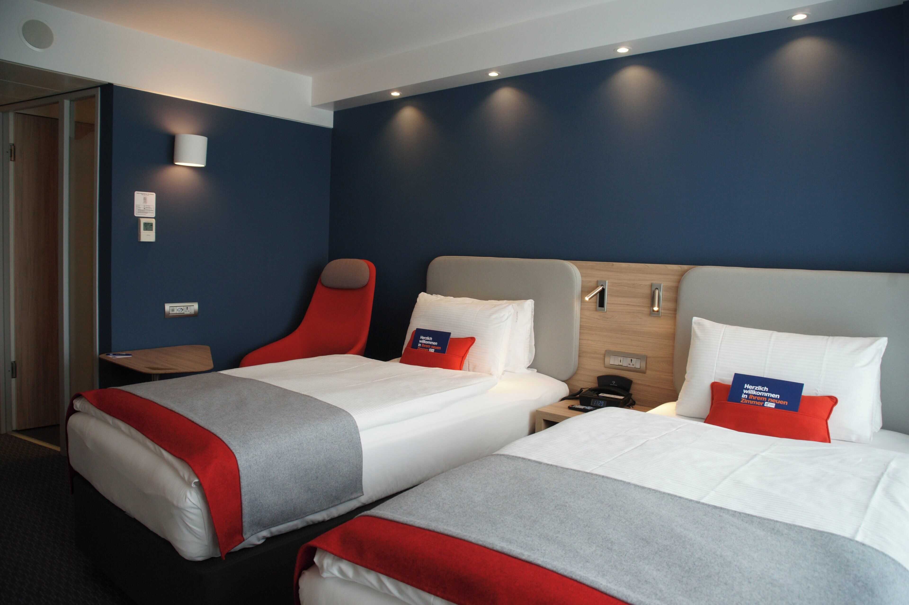 A hotel room with two beds, each with red and grey bedding, a wooden headboard, and a nightstand with a phone.