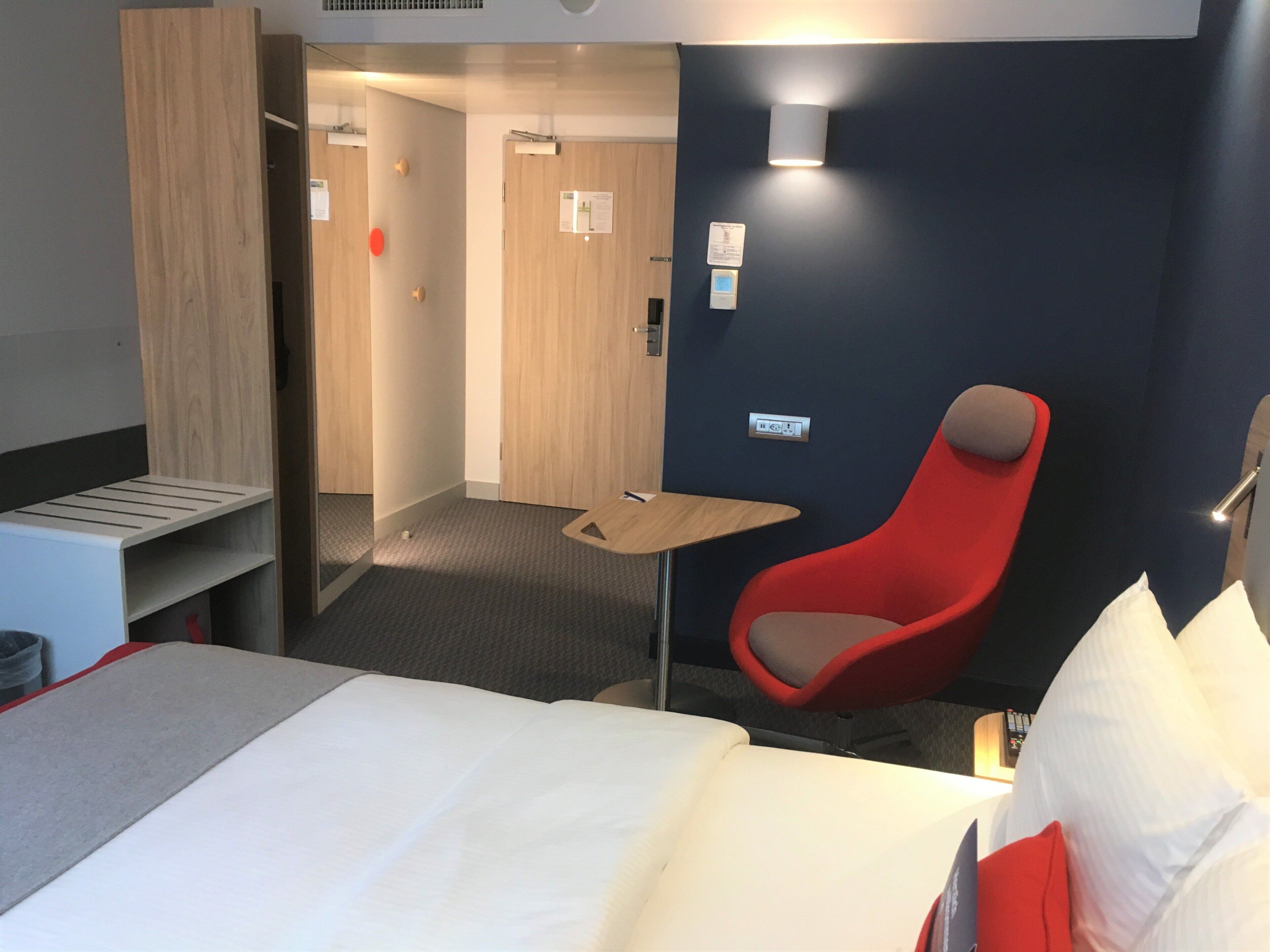 A hotel room with a bed, a red chair, a small table, and a wardrobe.