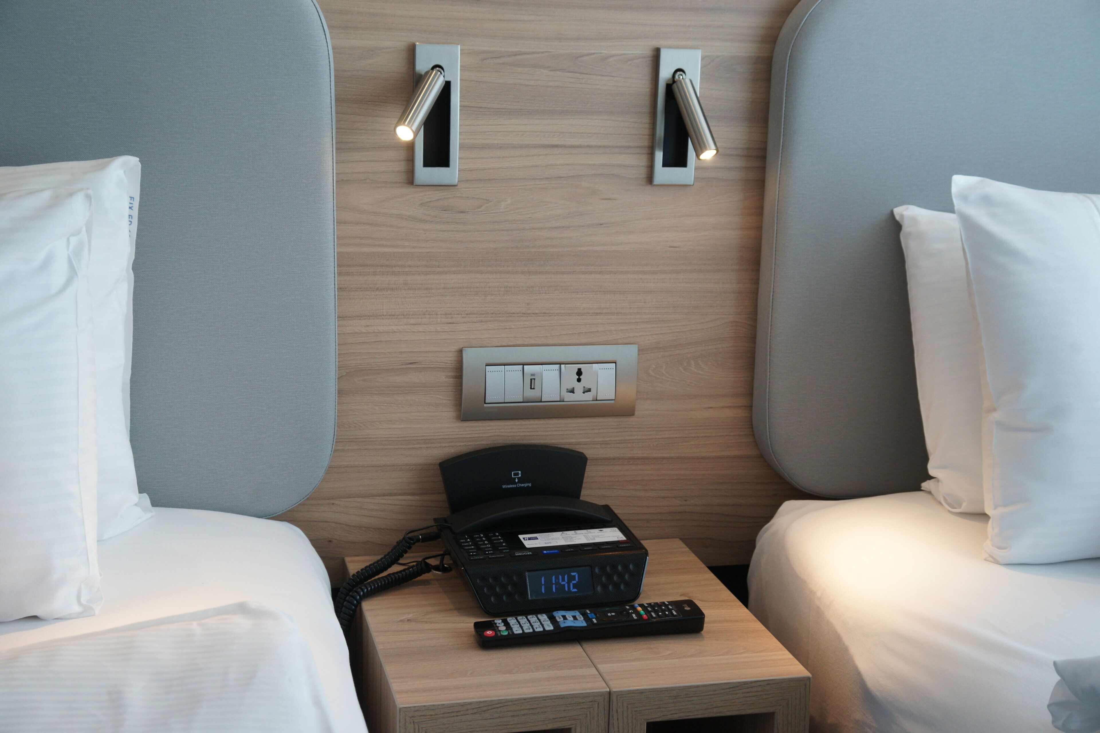 Habitación de hotel con dos camas, una mesita de noche con teléfono y controles remotos, y un enchufe en la pared.