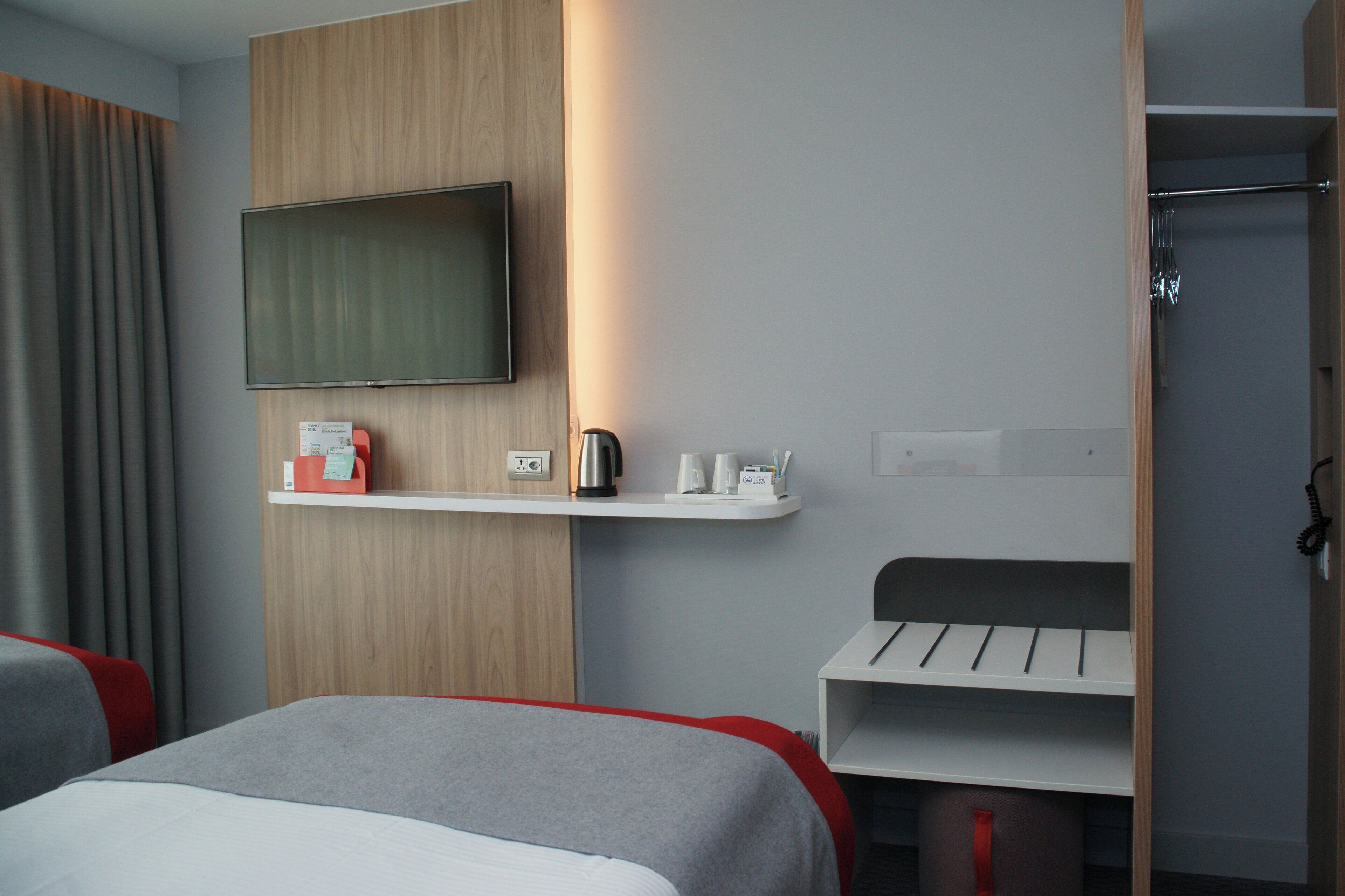 A hotel room with a bed, a TV mounted on the wall, a shelf with toiletries, and a wardrobe.