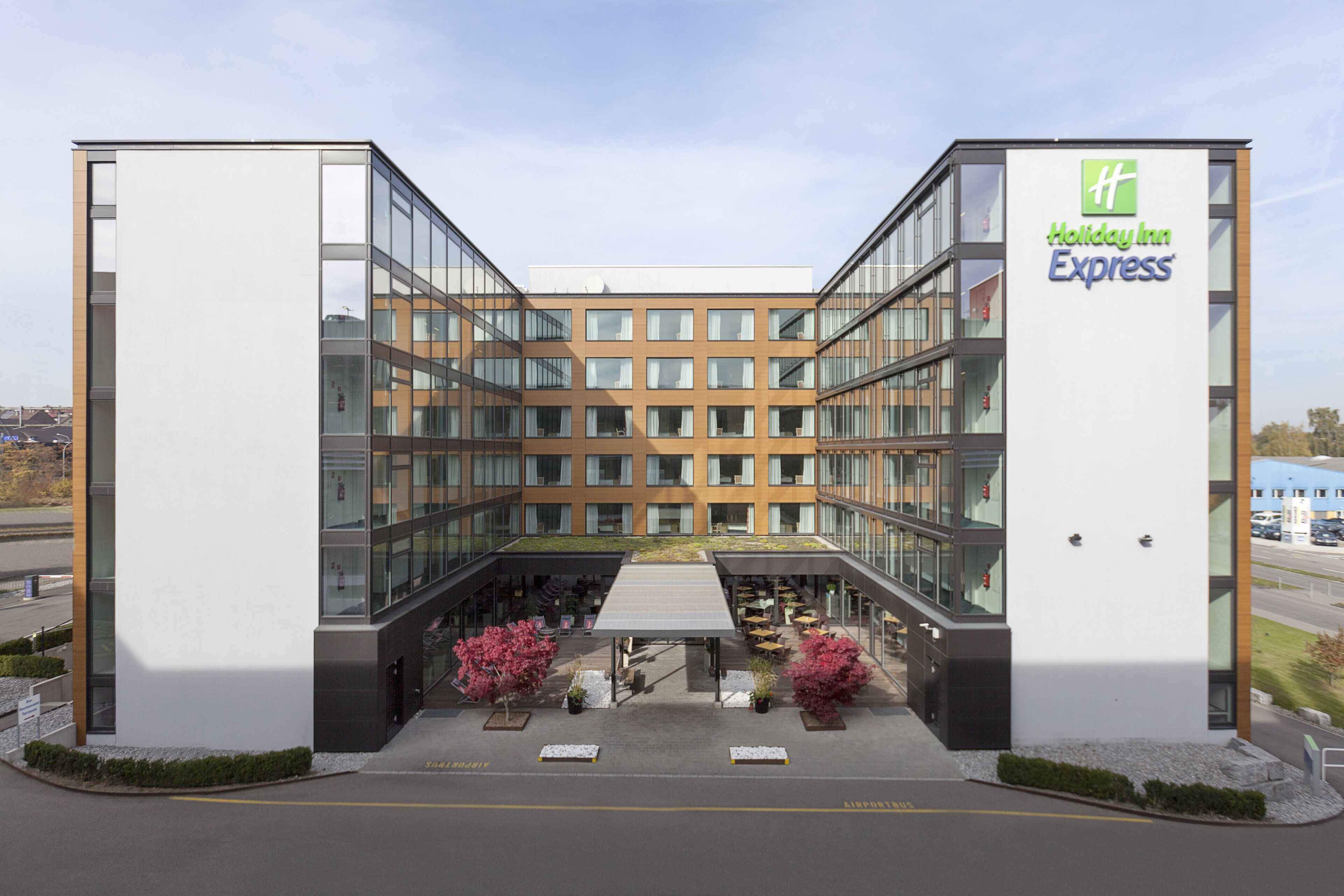 Et Holiday Inn Express hotel med en moderne glasfacade, udendørs siddeområde og et grønt tag.