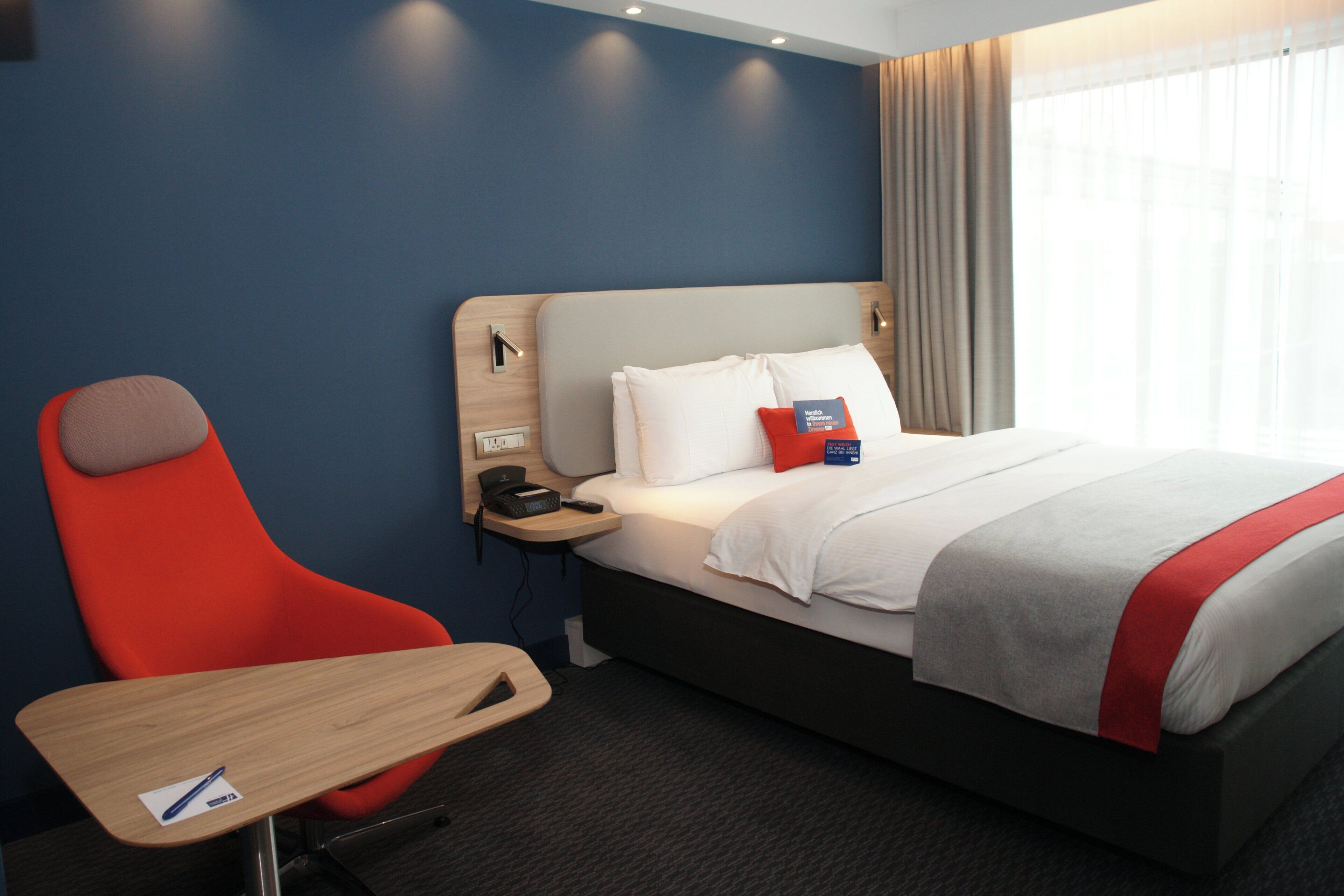 Habitación de hotel con cama, escritorio, silla, mesita de noche y ventana con cortinas.