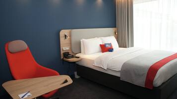 Quarto de hotel com cama, escrivaninha, cadeira, mesa de cabeceira e janela com cortinas.