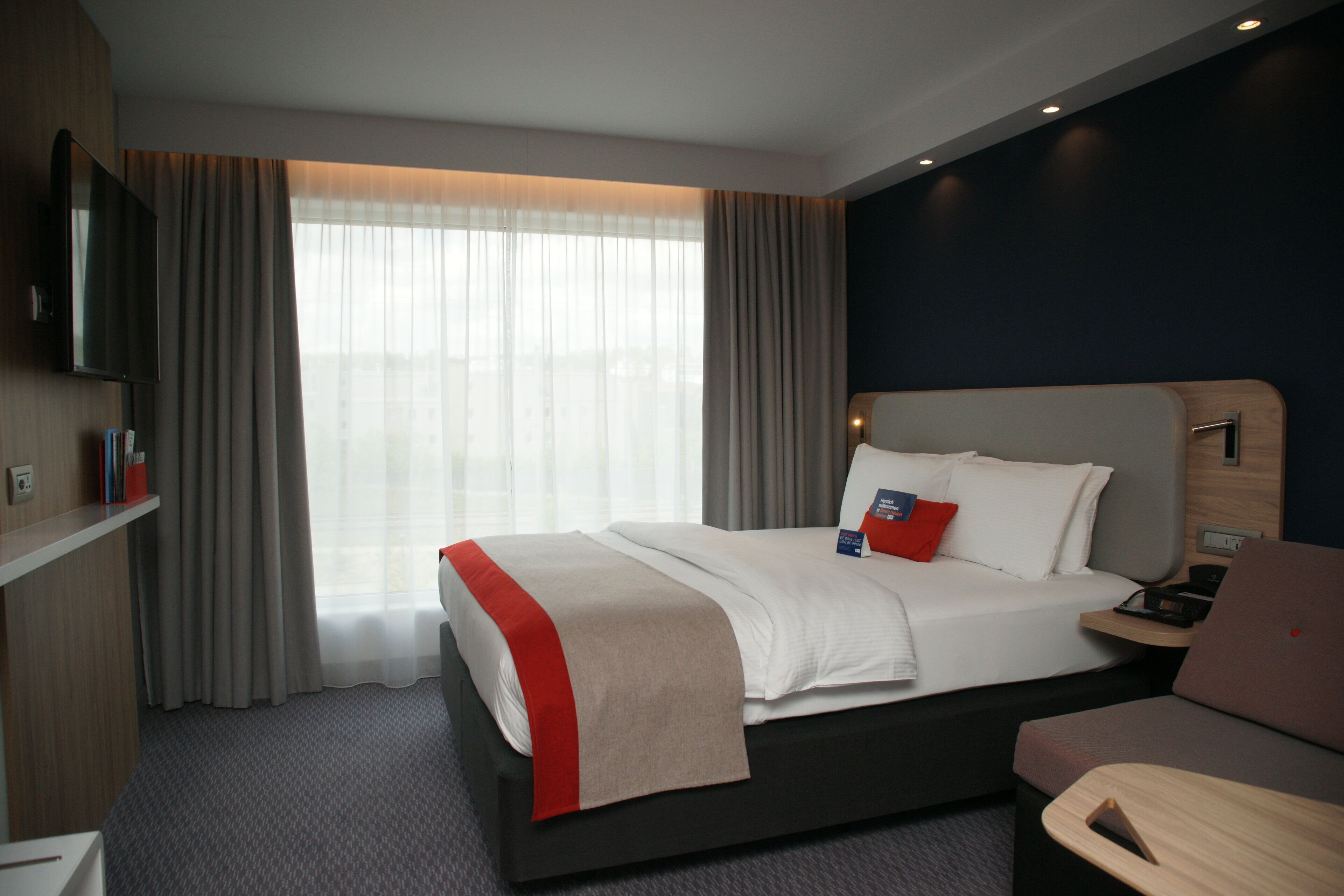 Habitación de hotel con cama, mesita de noche, una silla, televisor y ventana con cortinas.