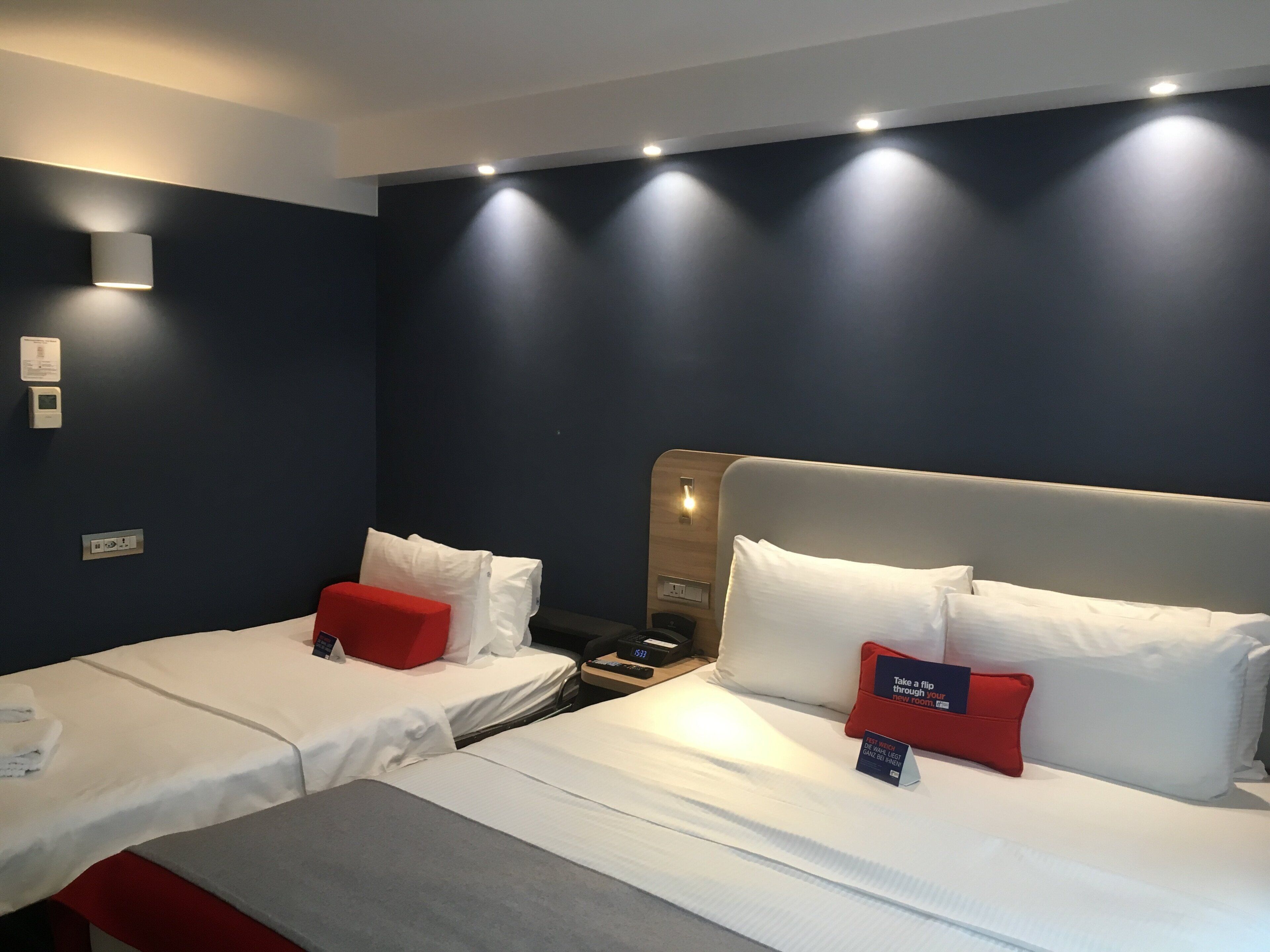 Una habitación de hotel con dos camas, una mesita de noche, un cojín rojo, un cartel y un termostato en la pared.