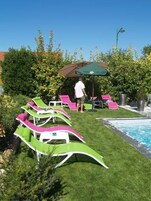 Zona de piscina con sillones reclinables verdes y rosados, un hombre caminando y una sombrilla verde.