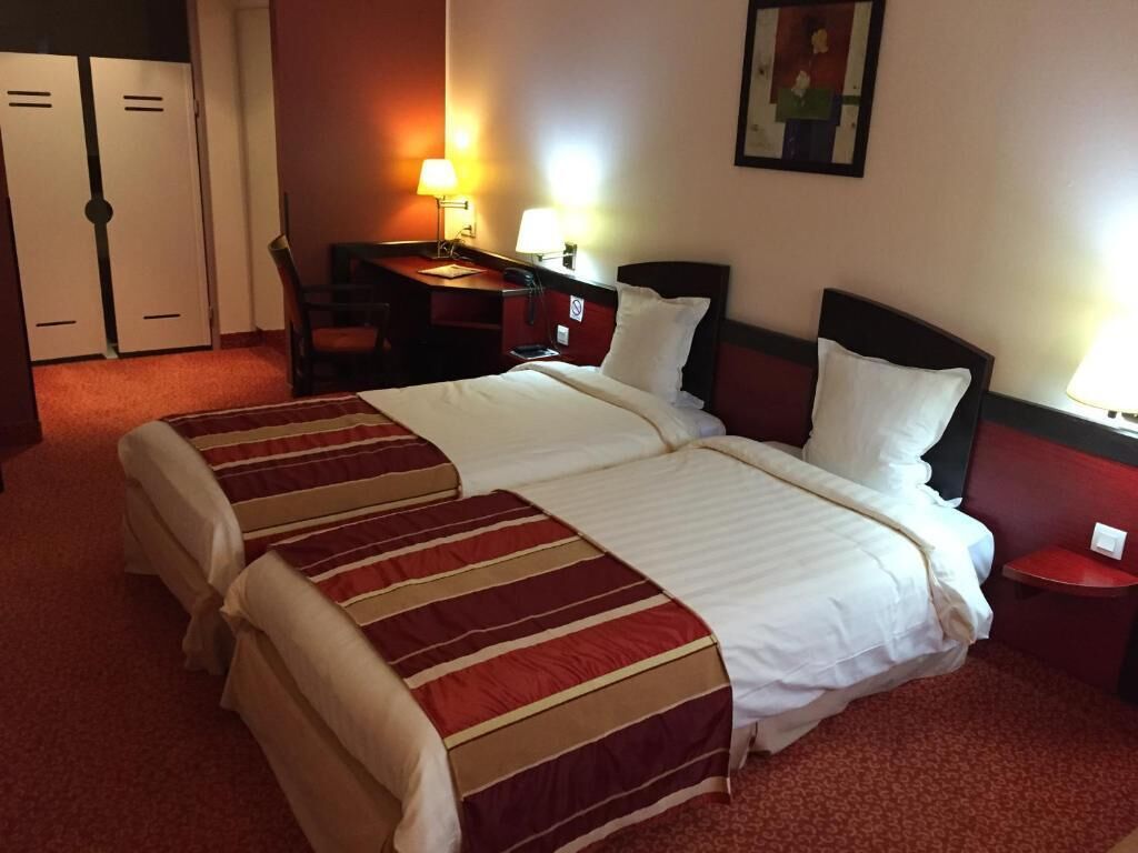 A hotel room with two beds, a desk, a chair, and a lamp.