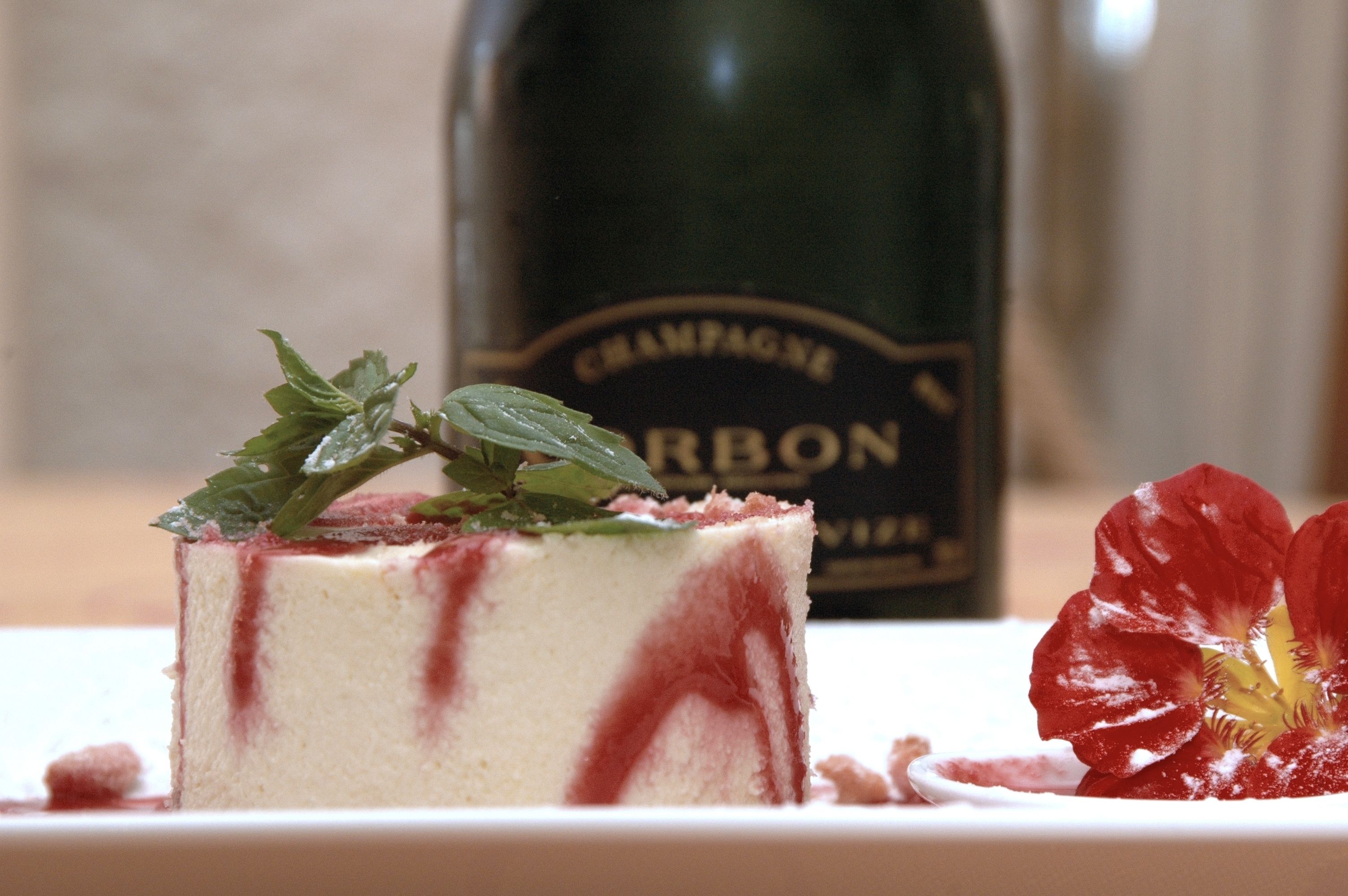 Une part de gâteau nappée d’une sauce aux fruits rouges et décorée de feuilles de menthe, une bouteille de champagne et une fleur rouge sur une assiette blanche.