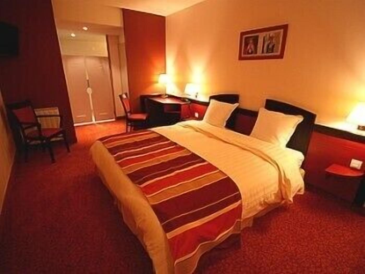 A hotel room with a large bed, a desk with a chair, a small table with a lamp, and a door leading to another room.