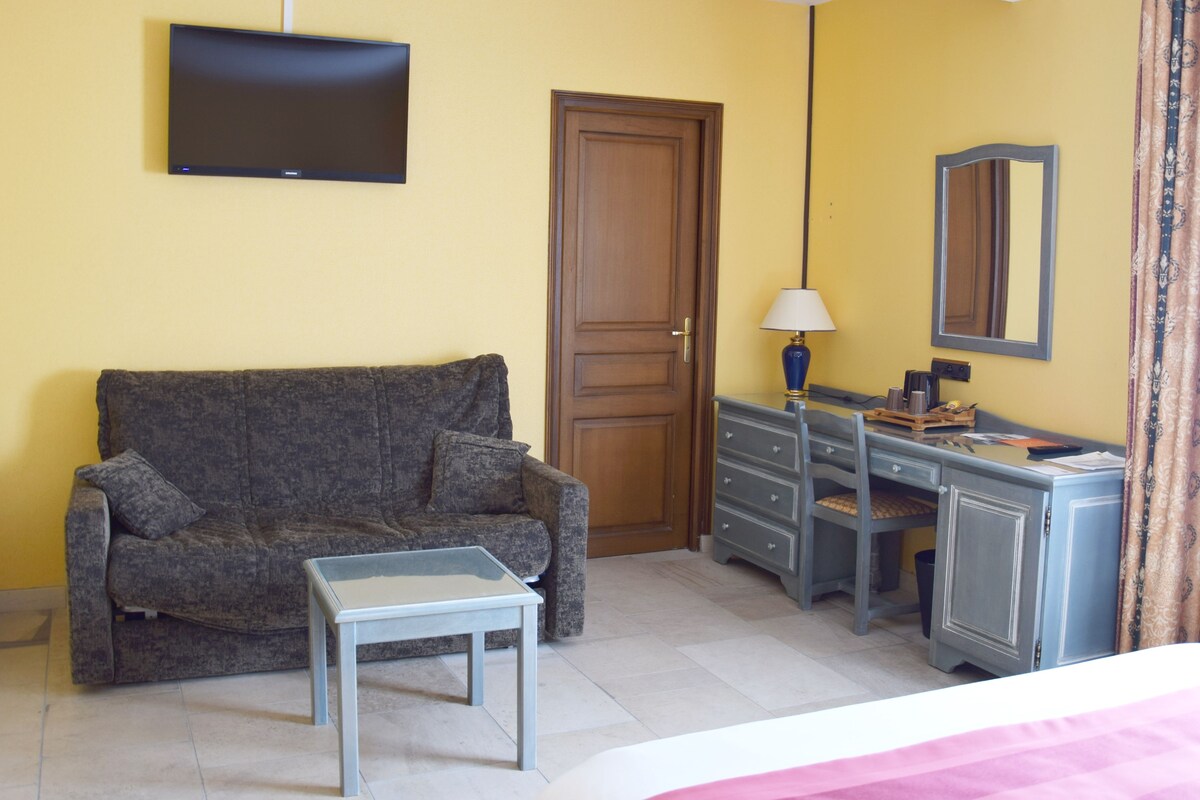A room with a sofa, a small table, a desk with drawers, a mirror, and a television mounted on the wall.