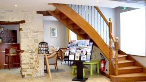 A hotel lobby with a wooden staircase, a stone wall, a reception desk, and a seating area with chairs.