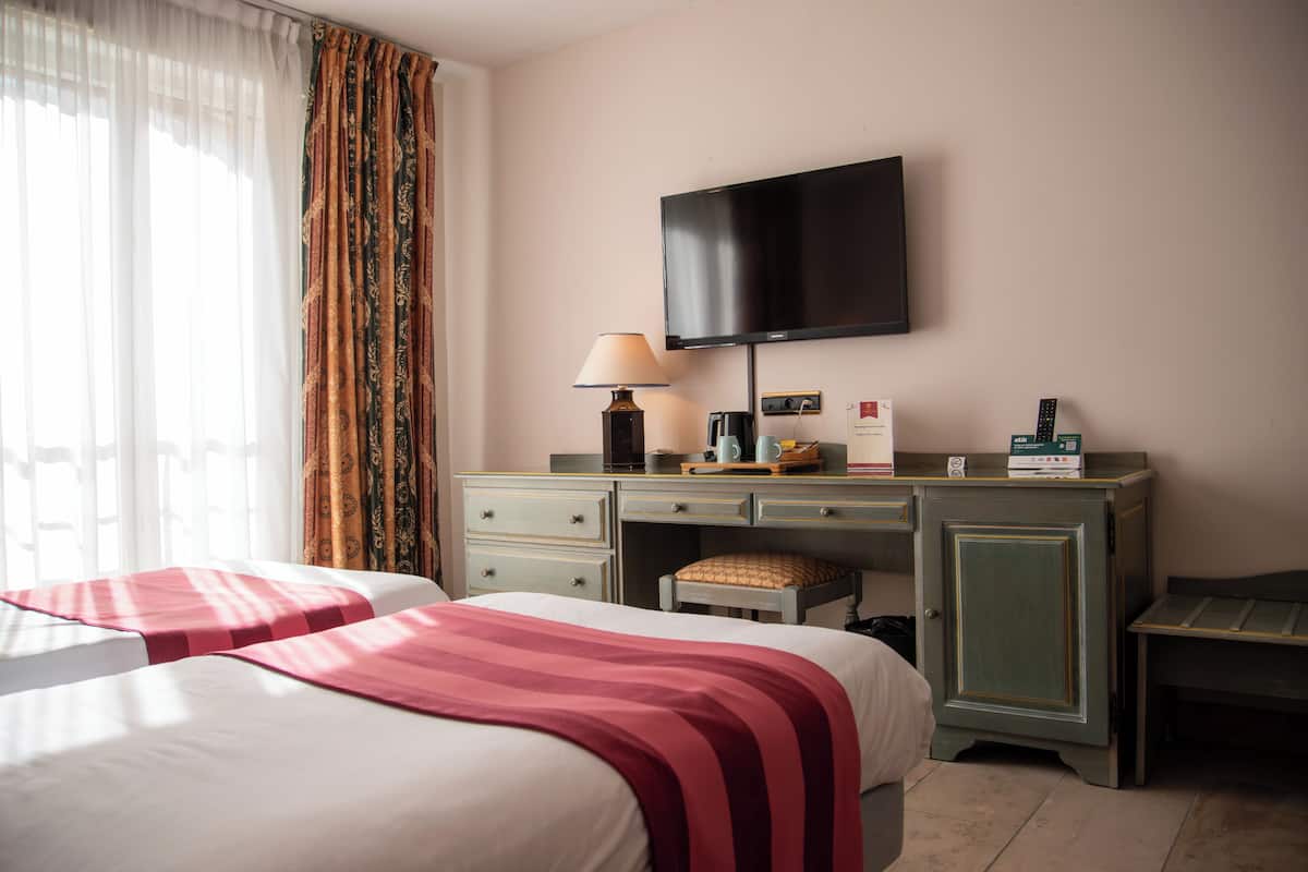 A hotel room with two beds, a desk with a television, a lamp, and a small table.