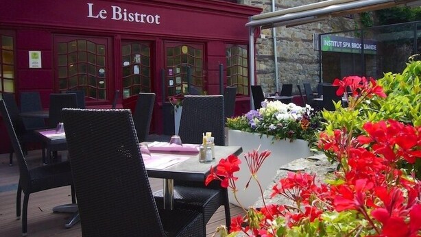 An outdoor seating area with tables and chairs, a stone building with 'Le Bistro' signage, and a flower bed with red flowers. - Hotel Le Relais De Broceliande (Paimpont)