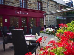An outdoor seating area with tables and chairs, a stone building with 'Le Bistro' signage, and a flower bed with red flowers. - Hotel Le Relais De Broceliande (Paimpont)