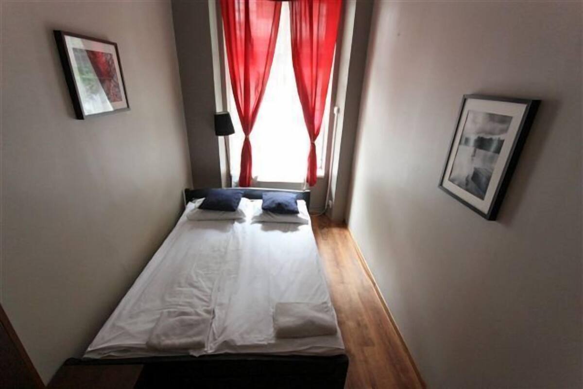 Una habitación pequeña con cama individual, ventana, cortinas rojas y dos cuadros en las paredes.