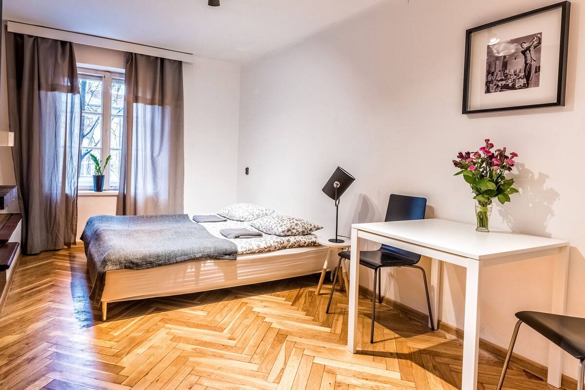Un dormitorio con suelo de madera en espiga, una cama con ropa de cama estampada, un escritorio con una silla, una planta en maceta y un cuadro en la pared.