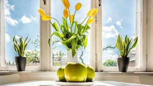 Una ventana con vista a un edificio, una mesa con una fuente de manzanas verdes y un jarrón con tulipanes amarillos.