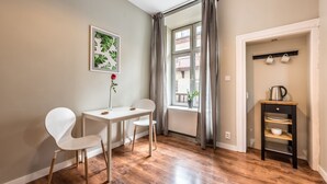 Una habitación pequeña y bien iluminada, con piso de madera, una mesa blanca con dos sillas, un cuadro en la pared, una ventana con cortinas y una pequeña zona de cocina con hervidor y tazas.