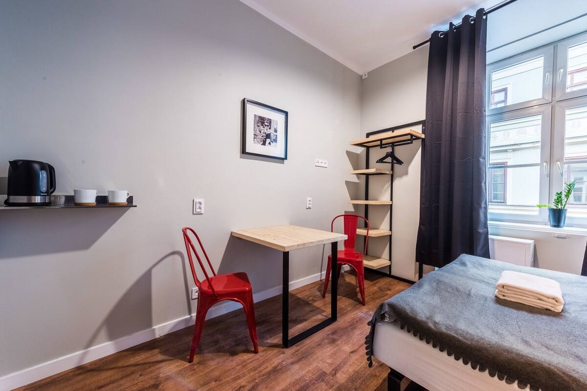 Una habitación pequeña y moderna con piso de madera, una mesa con dos sillas rojas, una cama con una colcha gris, una tetera negra sobre una repisa, un cuadro enmarcado en la pared y una ventana con cortina.