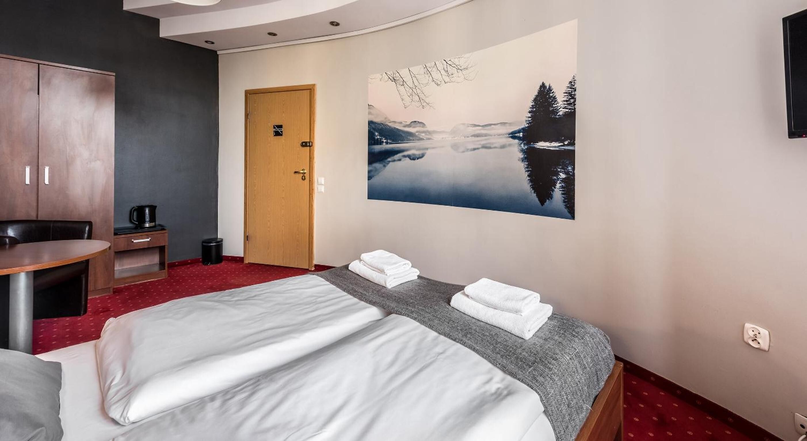 A hotel room with a bed, a desk, a wardrobe, and a large wall painting of a forest and lake.