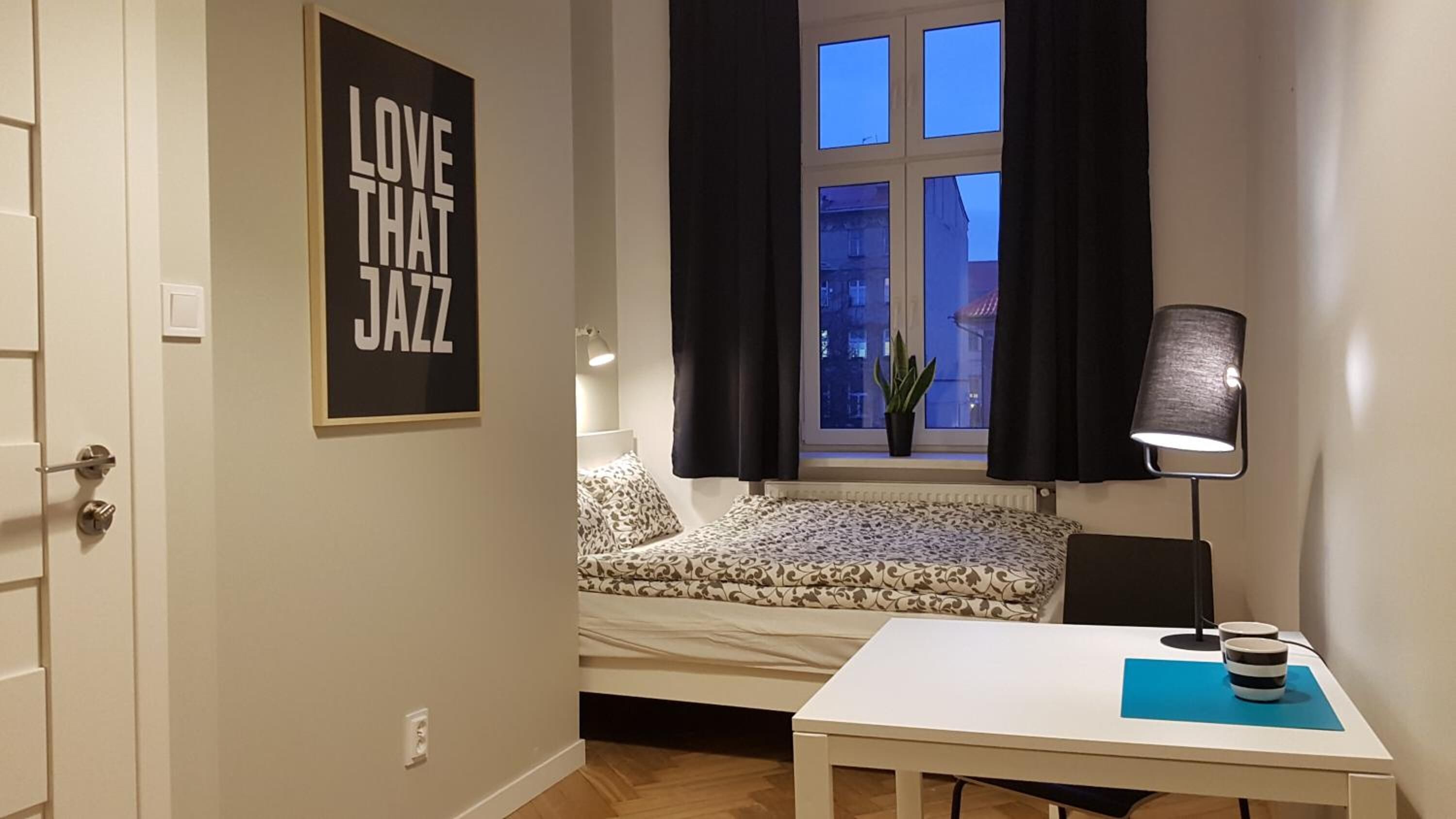 A small room with a bed, a table with a lamp, and a window with curtains.