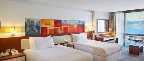 Un cuarto de hotel con dos camas, un escritorio, una silla, un ventanal con vista al mar y obras de arte en la pared.