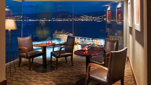 A room with a view of a cityscape at night, featuring a table and chairs, a lamp, and a window with artwork. - Kordon Hotel Pasaport (Izmir)