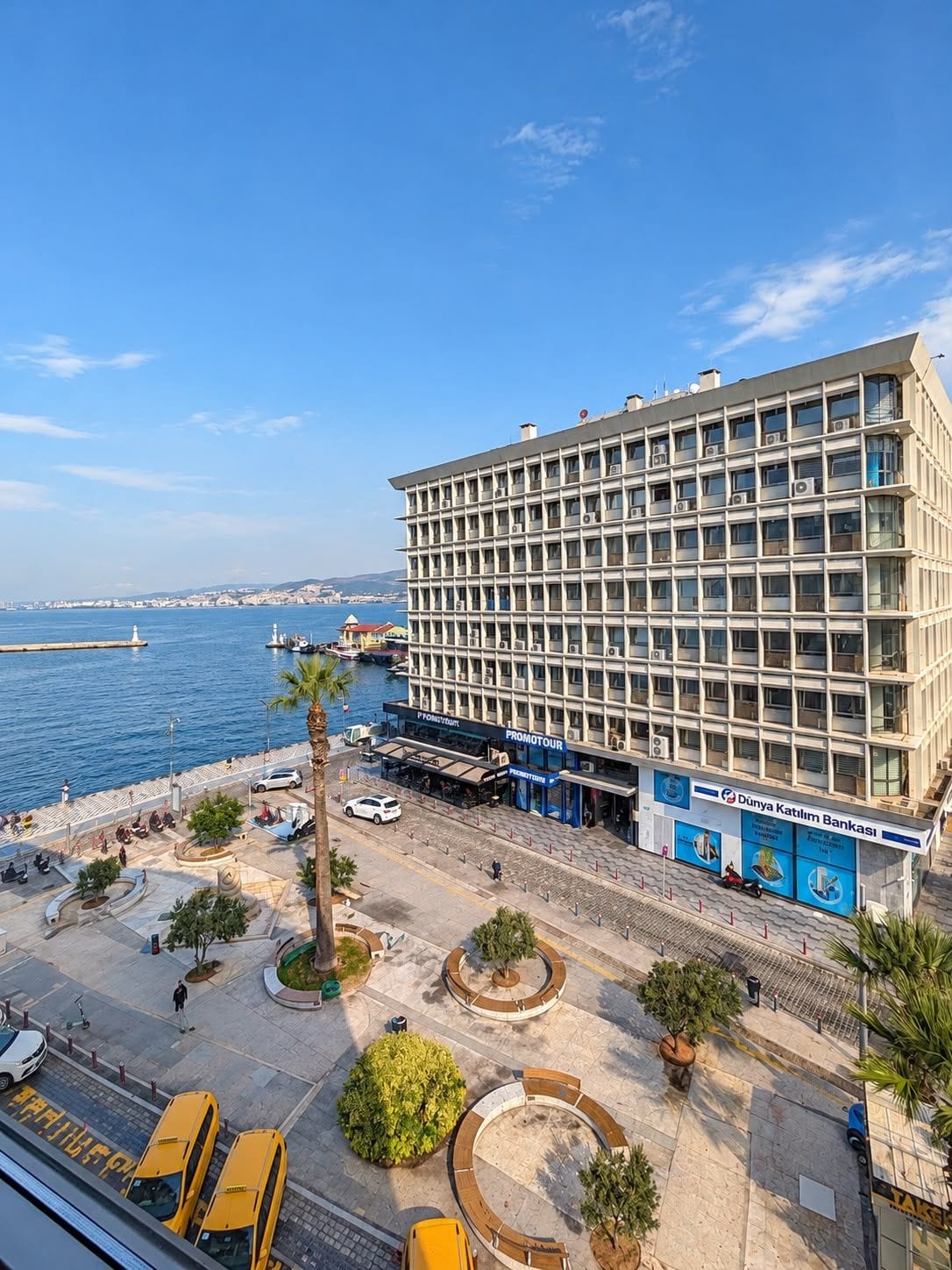 A multi-story hotel with a view of the sea, a parking area, and a promenade with trees and a fountain.