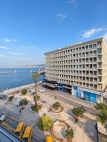 Un hotel de varios pisos con vistas al mar, un área de estacionamiento y un paseo marítimo con árboles y una fuente.