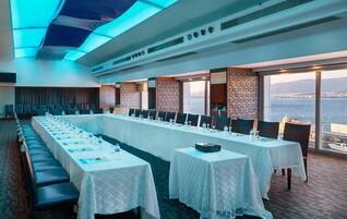 Un salón de conferencias con una larga mesa dispuesta para una reunión, amplias ventanas con vista al mar y un diseño de techo moderno.