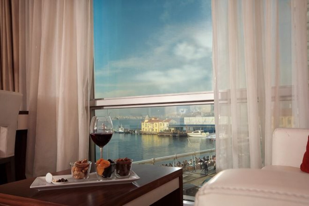 Quarto de hotel com uma grande janela para um corpo de água, uma mesa de jantar com um copo de vinho tinto, e vista para um edifício do outro lado da água.