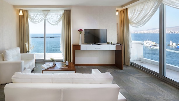 A hotel room with a large window offering a view of the sea and boats, a white sofa, a desk with a TV, and a small coffee table. - Kordon Hotel Pasaport (Izmir)