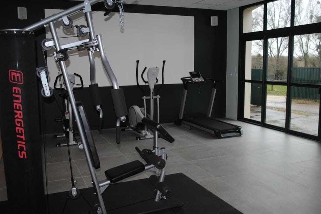 Academia com equipamentos de ginástica, esteira e uma grande janela.