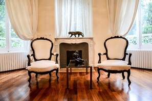 A room with two chairs, a fireplace, and a view of greenery through large windows. - Château de la Dame Blanche (Geneuille)