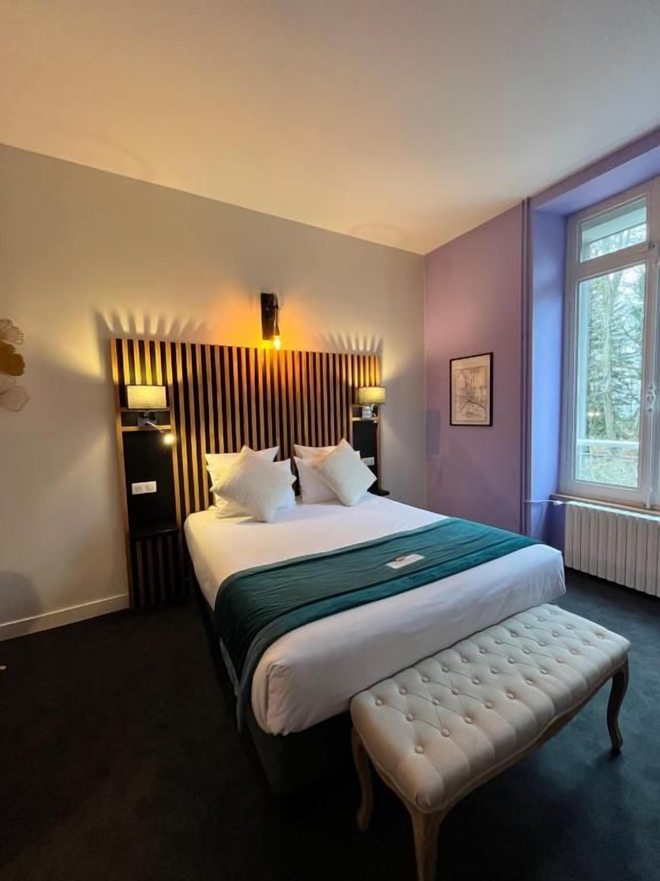 A hotel room with a bed, bedside tables, a bench, a window with a view, and a wall-mounted light fixture.