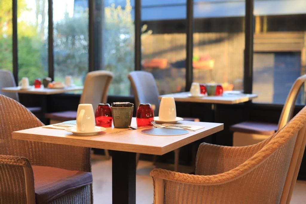 Un espace repas avec des tables préparées pour deux personnes, des chaises et une vue sur l’extérieur grâce à de grandes fenêtres.
