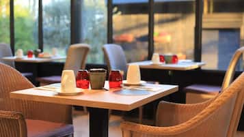 Un espace repas avec des tables préparées pour deux personnes, des chaises et une vue sur l’extérieur grâce à de grandes fenêtres.