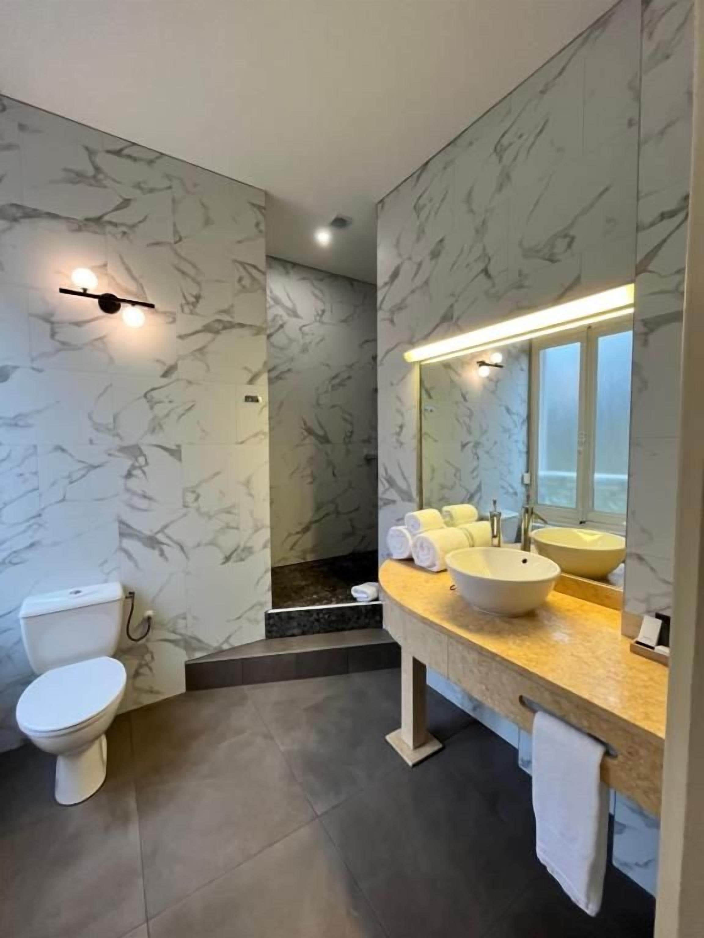 A modern bathroom with a marble wall, a toilet, a sink with a mirror, and a shower area.