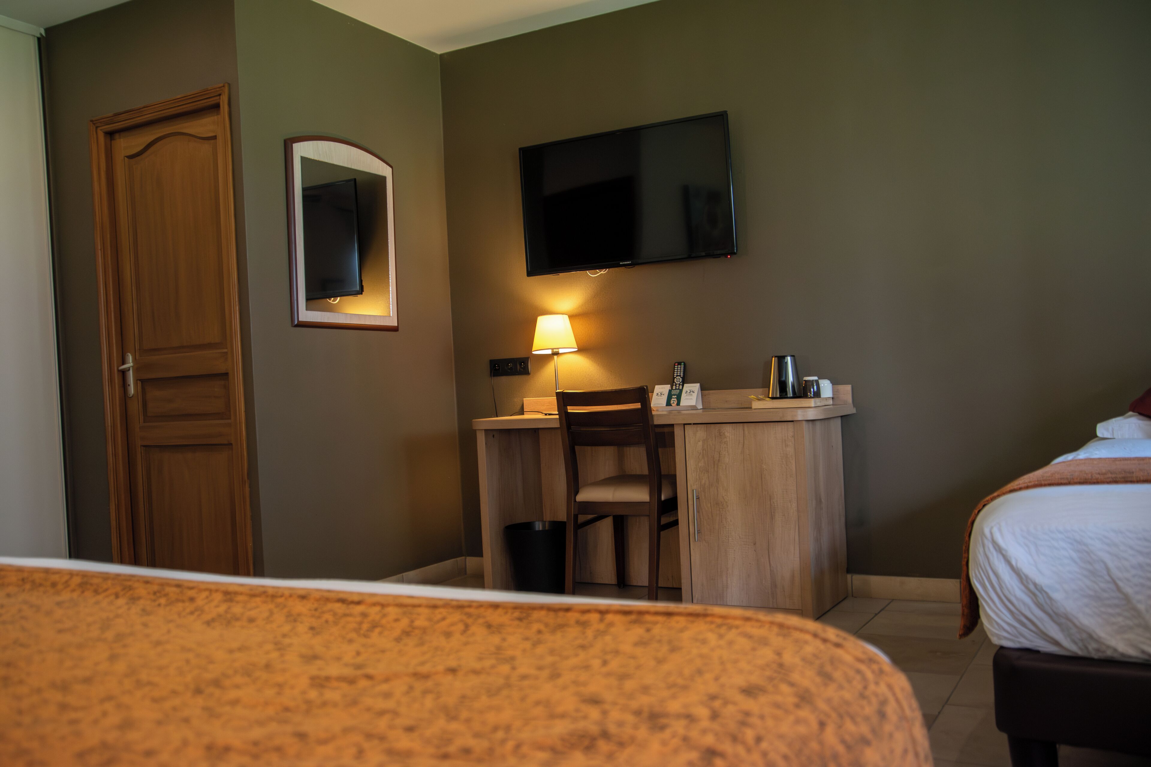 Une chambre d’hôtel avec un bureau en bois, un téléviseur fixé au mur, un lit recouvert d’une couette blanche et une petite table de chevet avec une lampe.