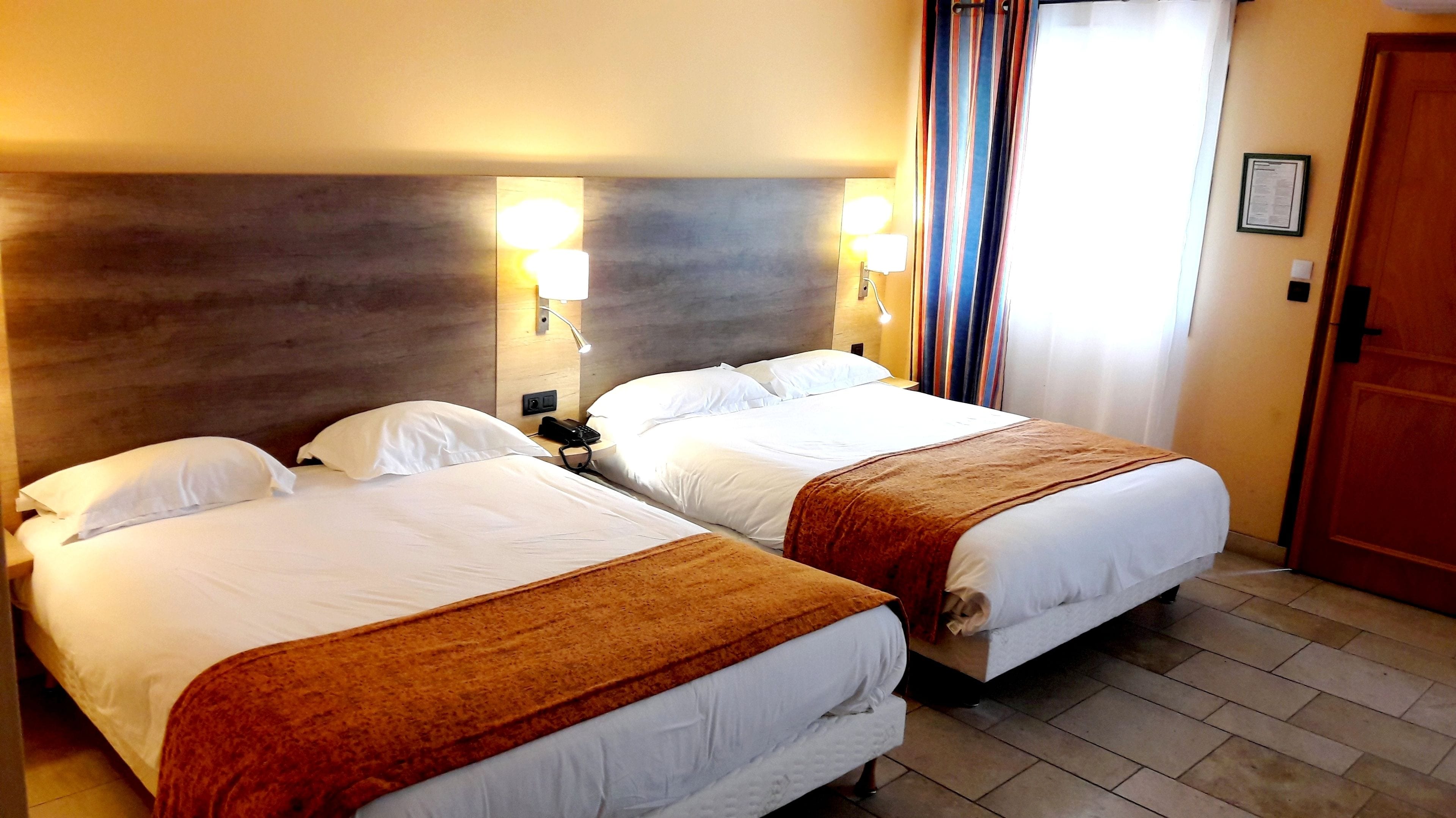 A hotel room with two beds, each with white linens and a brown throw blanket. There are wall-mounted lamps on either side of the beds, and a door with a wooden frame to the right.