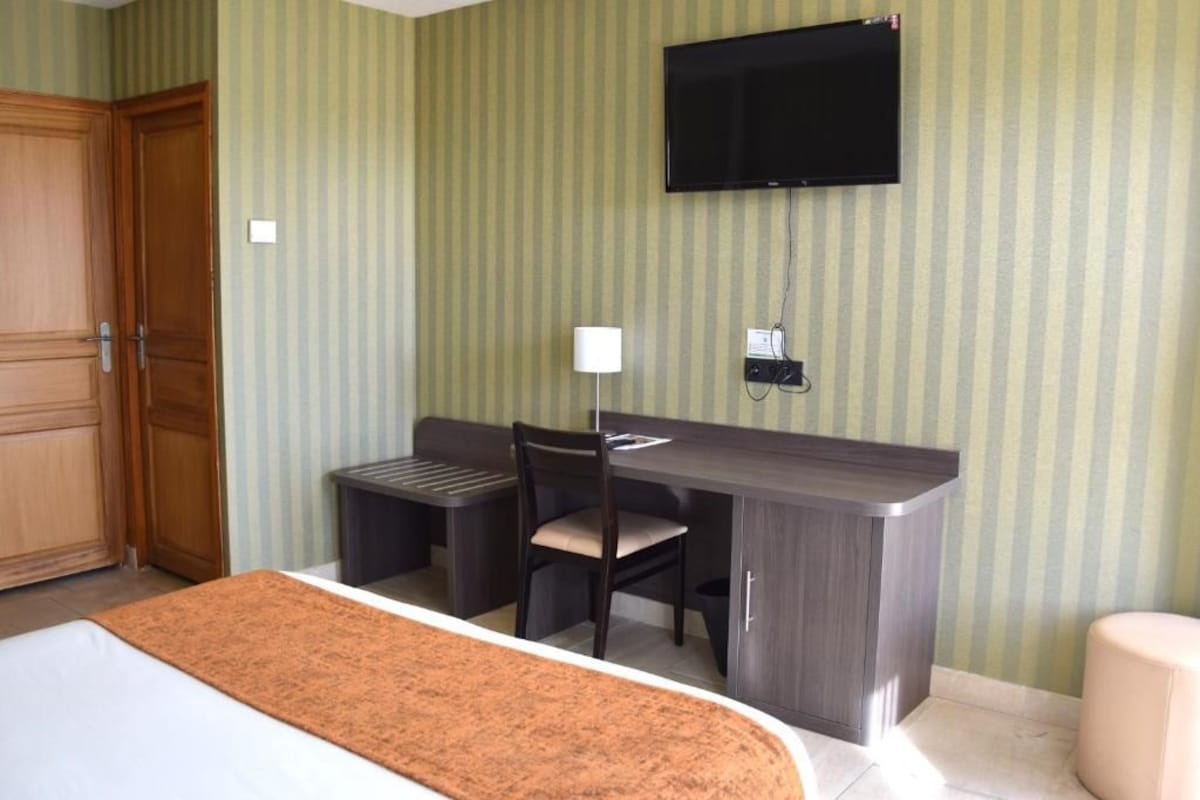 A hotel room with a bed, a desk with a chair, a TV mounted on the wall, and a small table with a lamp.