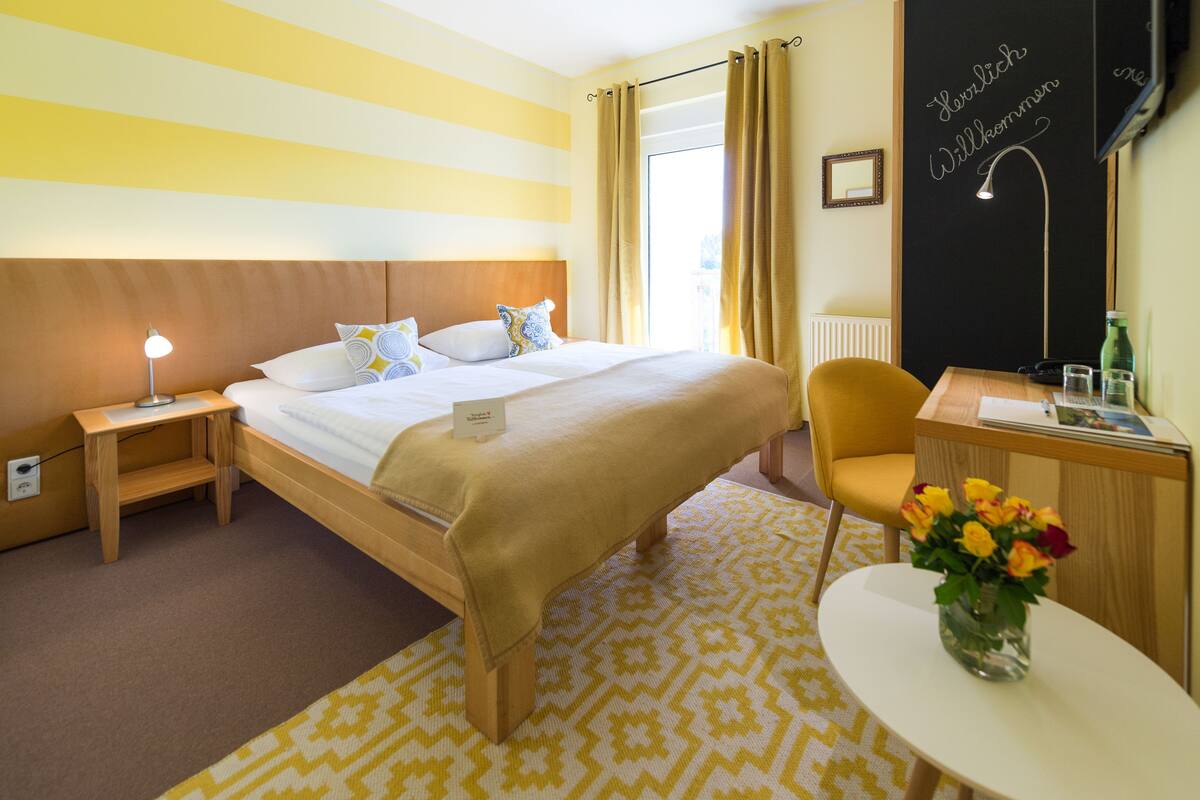 Et hotelværelse med en seng, et natbord, en stol, et skrivebord og et lille bord med en vase blomster.