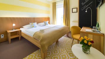 A hotel room with a bed, bedside table, chair, desk, and a small table with a vase of flowers.
